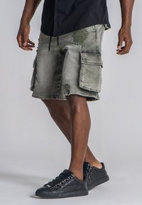 Pantalones cortos de carga en denim verde desgastado con parches desgastados, múltiples bolsillos laterales y un ajuste relajado. Combinados con zapatillas negras de corte bajo.