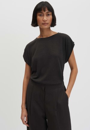 Femme aux cheveux noirs et lisses portant un haut noir ample à manches courtes et un pantalon noir taille haute, debout avec une main dans la poche.