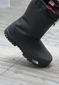 Bota negra impermeable con una superficie texturizada, un upper flexible y una suela gruesa y resistente. Logo notable en el lateral y parte superior acolchada.