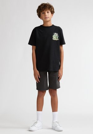 T-shirt en coton noir avec un motif graphique sur le côté gauche de la poitrine, associé à un short gris foncé. Des baskets blanches et des chaussettes complètent la tenue.