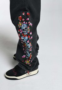 Jeans negros con bordados coloridos de flores y calaveras a lo largo del costado; combinados con zapatillas en blanco y negro con acentos rojos.