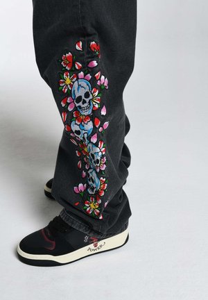 Black jeans with colorful floral and skull embroidery along the side; paired with black and white sneakers featuring red accents.