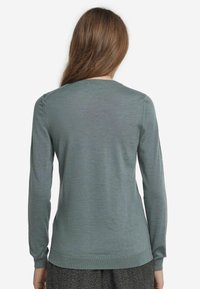 Kvinna med långt brunt hår som bär en enkel grå långärmad tröja och mörkgrå byxor, vänd bort mot en vit bakgrund.