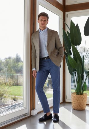 Hellbrauner Blazer, hellblaues Hemd, marineblaue Hose und schwarze Slipper. Glatte Stoffe, taillierte Passform und minimalistisches Design. Innenbereich.
