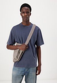 Sivi crossbody ruksak s patentnim zatvaračem, tkanim remenom; nosi se preko plave majice i izblijedjelih plavih traperica, na bijeloj pozadini.