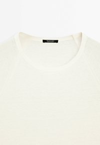 Cremefarvet, letvægts bomulds t-shirt med rund hals og Raglan-ærmer, der har en subtil tekstur og et svart mærke ved halsudskæringen.