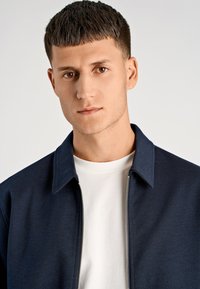 Granatowa kurtka zapinana na zamek z kołnierzem, wykonana z teksturowanej tkaniny, noszona na białej koszulce. Zbliżenie na górną część ciała i twarz.