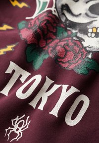 Il tessuto color marsala presenta una stampa grafica con un teschio, rose e fulmini, accentuata da un testo bianco screpolato che legge "TOKYO" e un ragno.