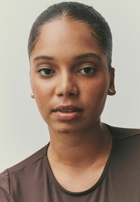 Záber na osobu so svetlohnedou pokožkou, ktorá má oblečenú tmavohnedú košeľu. Výrazné sú upravené vlasy, jemné náušnice a hladká pokožka.