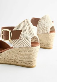 Sandali con zeppa e tomaia intrecciata in beige chiaro, cinturino marrone con fibbia dorata, e tacco in juta testurizzata.