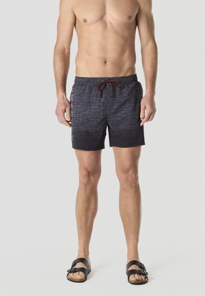 Hombre sin camisa vistiendo pantalones de baño negros con estampado y texto lateral, y sandalias negras de doble correa, de pie contra un fondo liso.