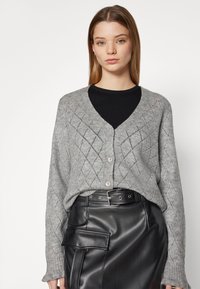 Gray knit cardigan with a diamond pattern, v-neck, and buttons. Worn over a black top, paired with a black leather skirt featuring pockets.