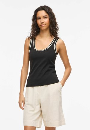 Jeune femme aux cheveux bouclés portant un haut sans manches noir avec un liseré blanc et un short beige arrivant aux genoux, debout avec une main dans la poche.