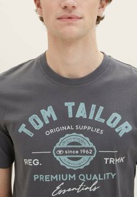 Graues Baumwoll-T-Shirt mit Rundhalsausschnitt und einem Frontgrafik in Hellblau und Weiß, das den Markennamen und einen Vintage-Stil Text zeigt.