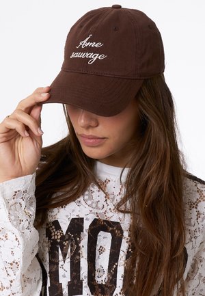 Bruine baseballcap met witte geborduurde tekst "âme sauvage." Heeft een ronde kroon en een gebogen klep, gemaakt van zachte stof.