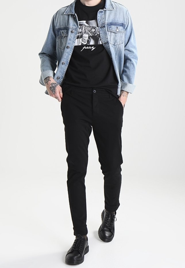 Denim takki, jossa vaalea pesu, musta graafinen t-paita, kapeat mustat housut ja mustat nauhakenkä. Ranteessa näkyvät tatuoinnit.