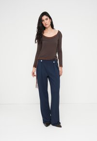 Καφέ μακρυμάνικο τοπ με στρογγυλή λαιμόκοψη, συνδυασμένο με σκούρες μπλε παντελόνες φαρδιάς γραμμής που διαθέτουν δύο ασημένια κουμπιά και πλαϊνές ζώνες.