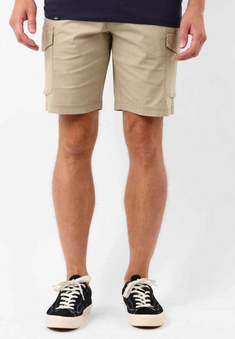 Shorts cargo beiges, longueur genou, en tissu mélange de coton, comportant des poches latérales, ourlet cousu, portés avec des baskets noires et des lacets blancs.
