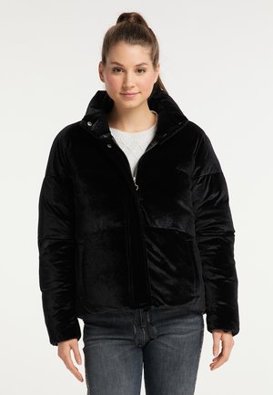 Junge Frau mit zurückgebundenen Haaren, die eine schwarze Steppjacke über einem weißen Strickpullover und grauen Jeans vor einem schlichten Hintergrund trägt.