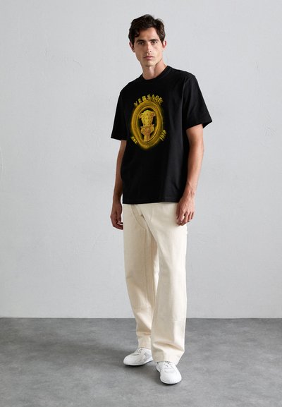 Man wearing black Versace T-shirt with golden Medusa logo, cream wide-leg pants, and white sneakers standing against a plain gray wall.