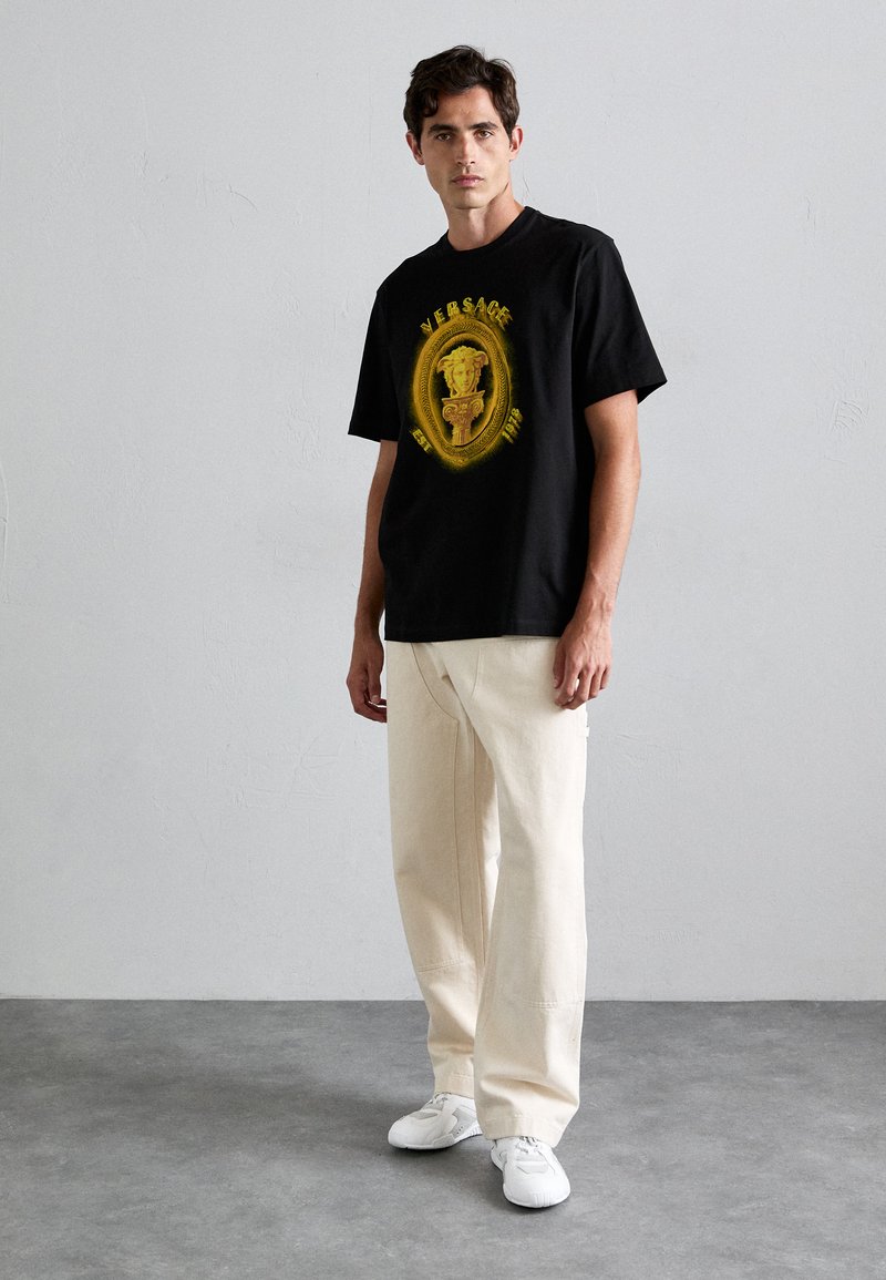 Man in een zwart Versace T-shirt met gouden Medusa-logo, crèmekleurige wijde broek en witte sneakers, staande tegen een effen grijze muur.