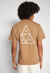 Camiseta de algodón marrón con un gran diseño geométrico triangular blanco donde se encuentran las letras H, U y F dentro de las formas.