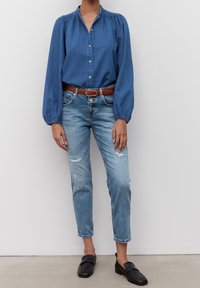 Camisa de mezclilla azul oscuro con botones en la parte delantera y mangas abullonadas, combinada con jeans desgastados azul claros y un cinturón marrón, finalizada con mocasines negros.