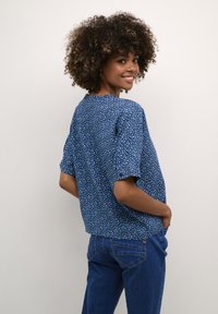 Mėlyna gėlėmis raštuota trumpa rankovėms bluzelė, pagaminta iš lengvos medžiagos, derinama su mėlynais džinsais, turinčiais užpakalinių kišenių detales.