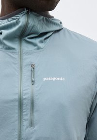 Άνδρας που φοράει ανοιχτό μπλε ζακέτα Patagonia με φερμουάρ, ψηλό γιακά και μικρή θήκη στο στήθος με φερμουάρ στην αριστερή πλευρά.