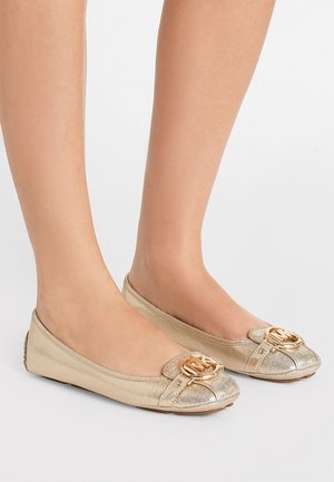 Femme portant des chaussures plates métalliques dorées avec un bout arrondi et un embleme décoratif doré sur le dessus, se tenant sur une surface blanche.