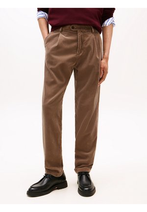 Homme portant un pantalon en velours côtelé marron et des chaussures en cuir noir, les mains dans les poches et un pull bordeaux laissant apparaître des manches de chemise rayée retroussées.