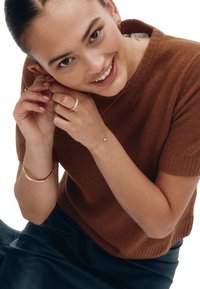 Brun stickad tröja med korta ärmar, guldsmycken inklusive hoops, ringar och ett armband. Person som justerar örhänge med ett leende.