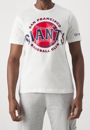 Mand iført hvid San Francisco Giants baseballklub T-shirt og lysegrå bukser står mod en ensfarvet baggrund.
