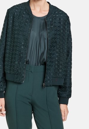 Bombers vert foncé ouvert sur le devant avec motif géométrique découpé sur un chemisier plissé vert foncé et un pantalon taille haute vert foncé.