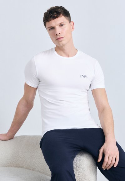 Νέος άνδρας με κοντά σγουρά μαλλιά, φοράει λευκό εφαρμοστό t-shirt και σκούρο παντελόνι, κάθεται σε καρέκλα με ανοιχτόχρωμη υφή.