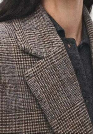 Gros plan sur un blazer à carreaux marron et gris porté sur une chemise gris foncé boutonnée, sur une personne aux longs cheveux foncés.