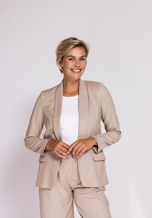 Smiling woman with short blonde hair wearing a beige suit and white top standing against a plain light background.
