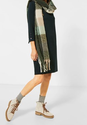 Mujer vistiendo un vestido negro hasta la rodilla, una bufanda larga de cuadros multicolor, calcetines grises y botas beige con cordones hasta el tobillo.
