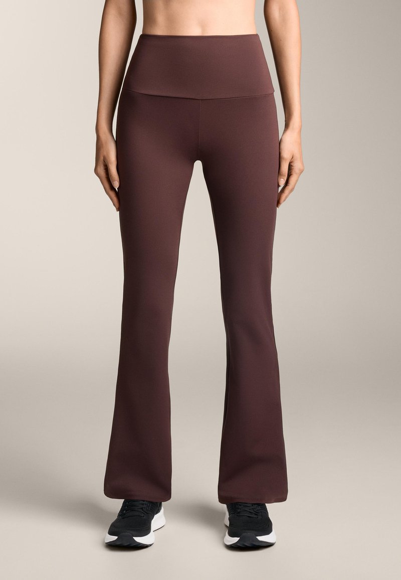 Legginsy z wysokim stanem i rozszerzanym dołem w ciemnym burgundowym kolorze, charakteryzujące się gładką teksturą i bezszwowym designem. W zestawie z czarnymi butami sportowymi.