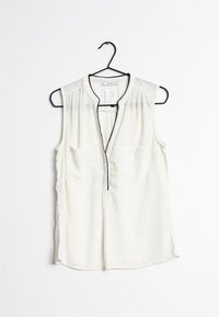 Sleeveless, cream blouse with a V-neck and black trim. Features two chest pockets and a lightweight, smooth texture. Hangs from a black hook.