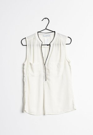 Sleeveless, cream blouse with a V-neck and black trim. Features two chest pockets and a lightweight, smooth texture. Hangs from a black hook.