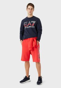 Felpa navy con testo bianco e rosso, abbinata a pantaloni corti rossi con branding bianco. Il modello indossa scarpe da ginnastica navy.