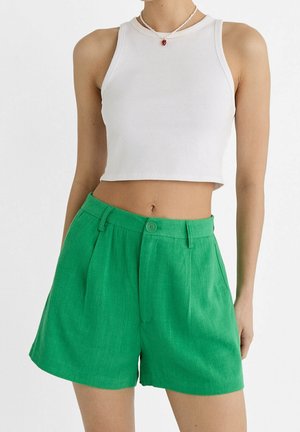 Femme portant un haut court blanc sans manches, un short vert taille haute et un collier de perles avec un pendentif rouge.