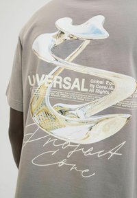 Pelēks T-krekls ar lielu grafiku, kas attēlo spīdīgu abstraktu spirālveida dizainu, ar tekstu "UNIVERSAL" un "Project Cone" kontrastējošā baltā krāsā.