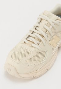 Sneaker beige con pannelli in rete, dettagli in pelle liscia, lacci testurizzati e un logo visibile sul lato.