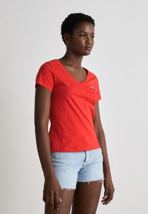T-shirt rossa in cotone con scollatura a V, maniche corte e design aderente. Presenta un piccolo logo ricamato sul davanti. Indossato con pantaloncini in denim chiaro.