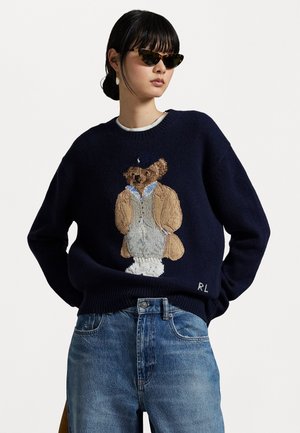 Suéter navy que presenta un intrincado diseño de oso tejido, con un pelaje marrón claro y camisa azul. Cuello acanalado y textura suave, combinado con jeans de mezclilla.