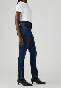 Tmavě modré pružné džíny s vysokým pasem, bočním zipem na lemu, které jsou kombinované s upnutým bílým tričkem a černými špičatými kotníkovými botami.