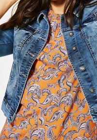 Jeansjacka med slitna detaljer, ljusblå färg och silverknappar som bärs med en orange klänning med lila paisleymönster.