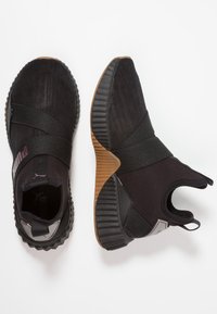 Svarta slip-on träningsskor med ribbade bruna och svarta sulor, elastiska korsremmar och Puma-logotyp på tungan och sidan av ena skon.